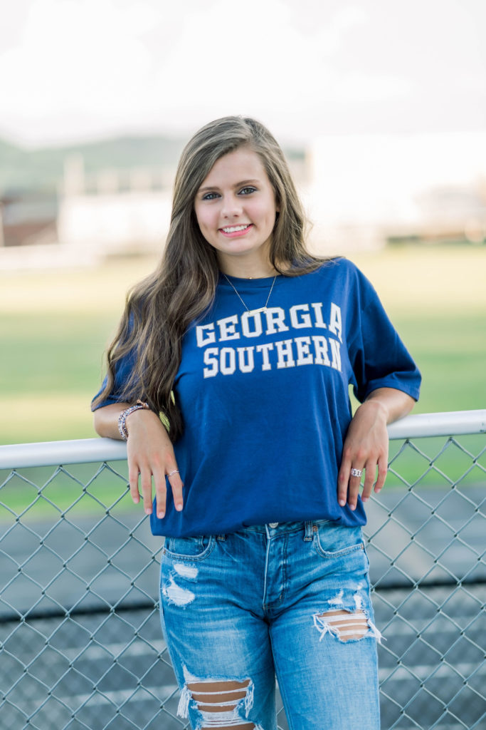 young woman in a Georgia Southern college t-shirt leaning on a chain link fence on a football fiel smiling at the camera for her senior photos