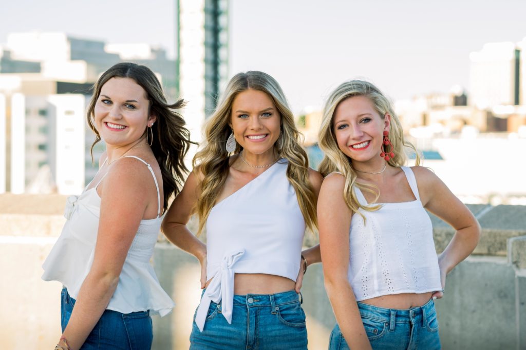 group senior sessionon top of a parking garage in Knoxville Tennessee. Seniors smiling with city scapr ein the background while wearing white tops and jeans. 