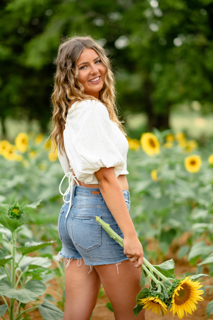 Young Tennessee woman wearing denim shorts, and white crop top with puffy sleeves holding sunflower during senior session