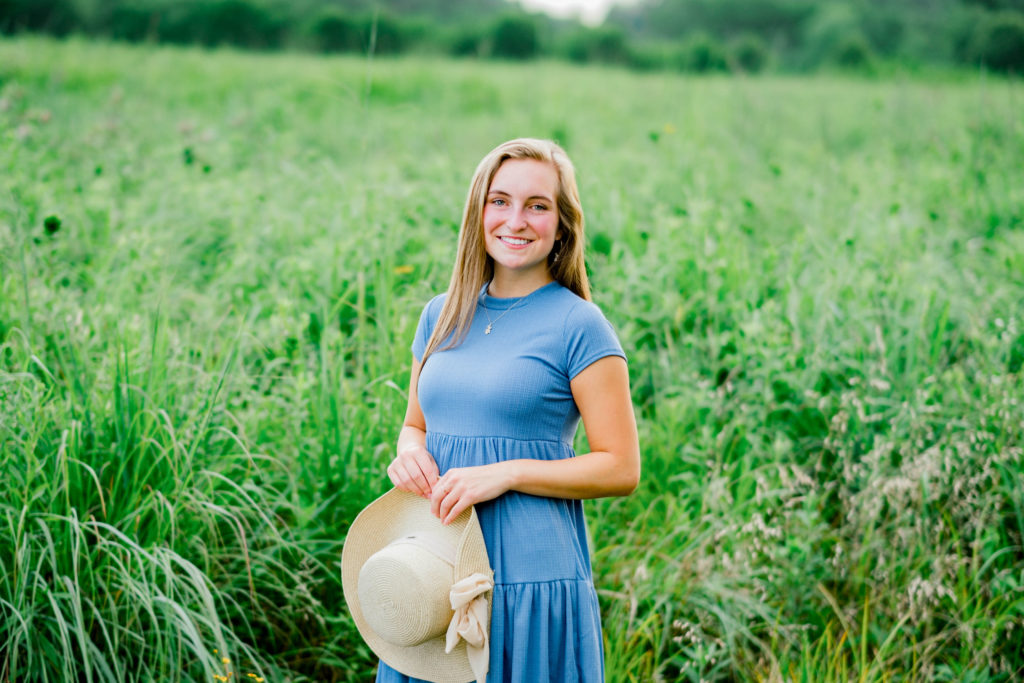 senior portrait fall outfit ideas with girl in a green field in Tennessee in a blue babydoll dress holding a hat and smiling at the camera for her senior photos