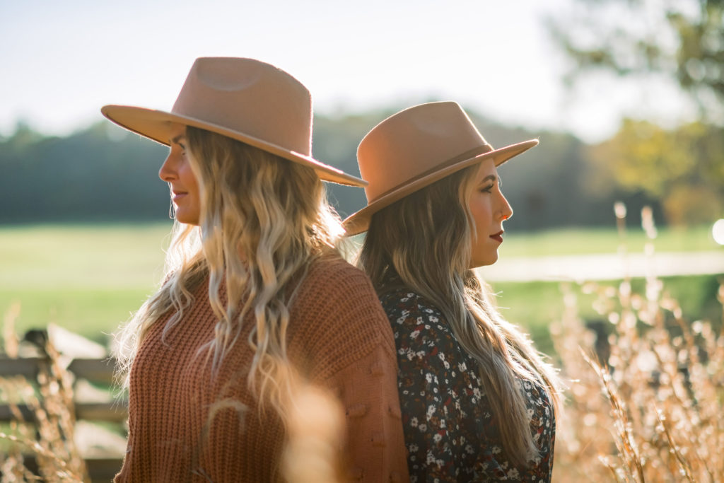 fall senior portrait outfit trend for girls with two seniors wearing wide brimmed camal fadora hats standing back to back in a field in the fall in Knoxville Tennessee
