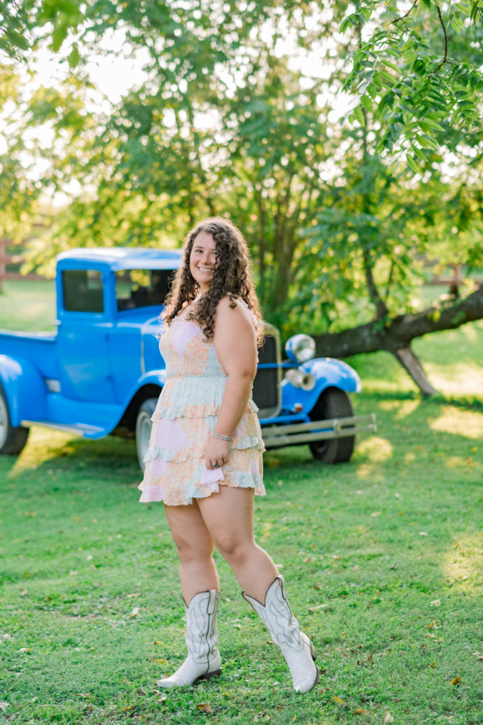 Jenna wearing fun dress and white cowboy boots in front of Grandpa's old blue pickup truck during outdoor session